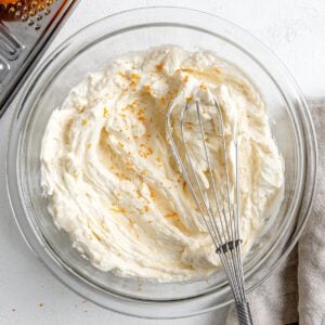 completed Orange Buttercream Frosting in a glass bowl with a whisk against a white background