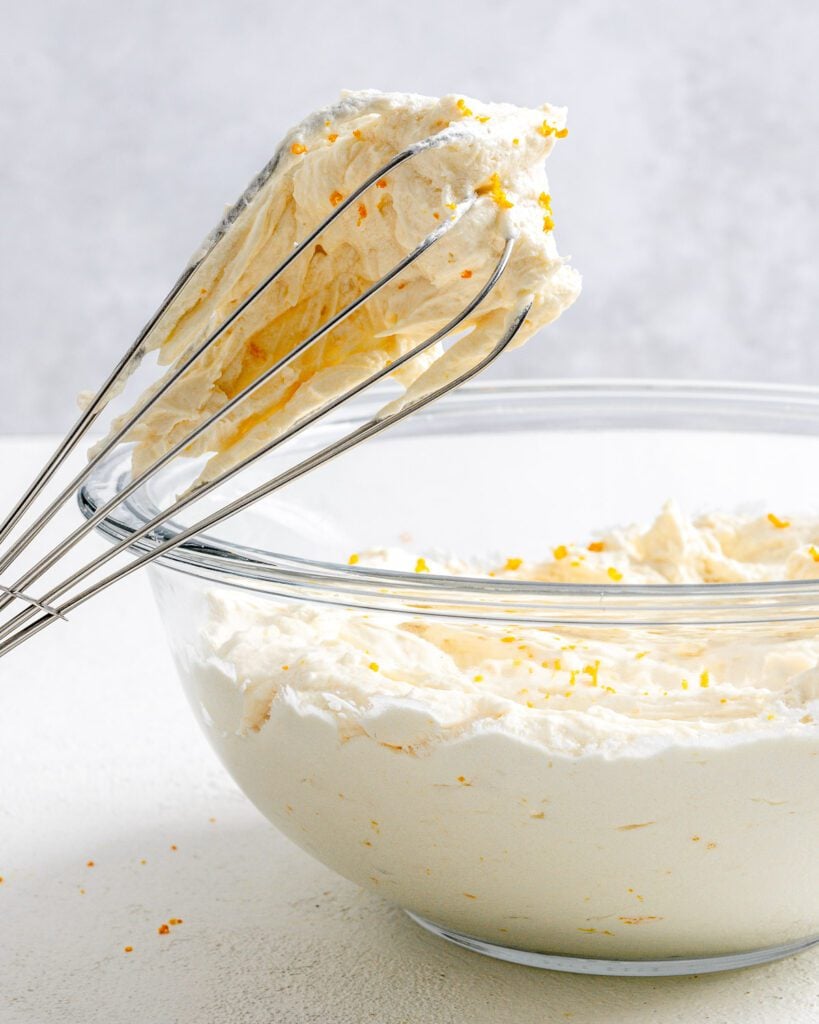 completed Orange Buttercream Frosting in a glass bowl with a whisk against a white background