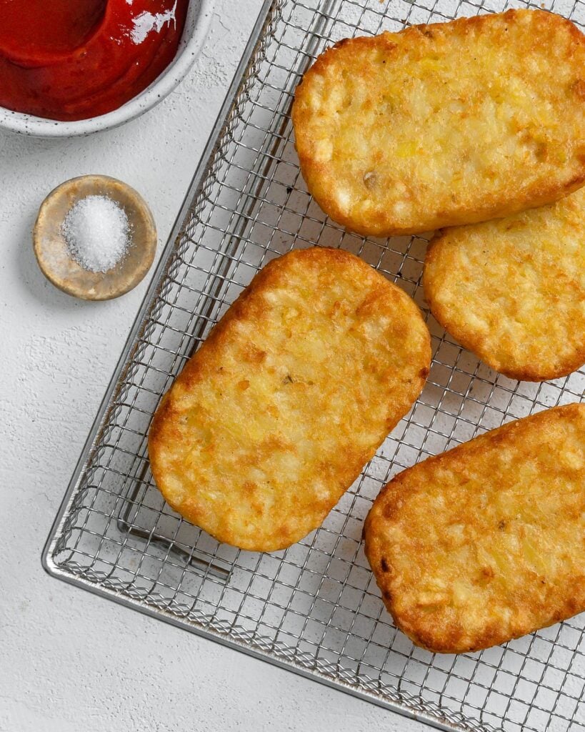 four completed hash brown patties on a air fryer tray with ketchup and salt in the background