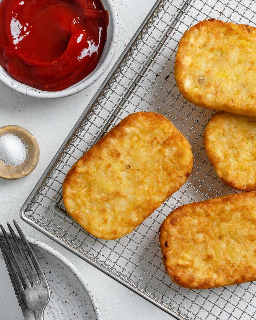 four completed hash brown patties on a air fryer tray with ketchup and salt in the background