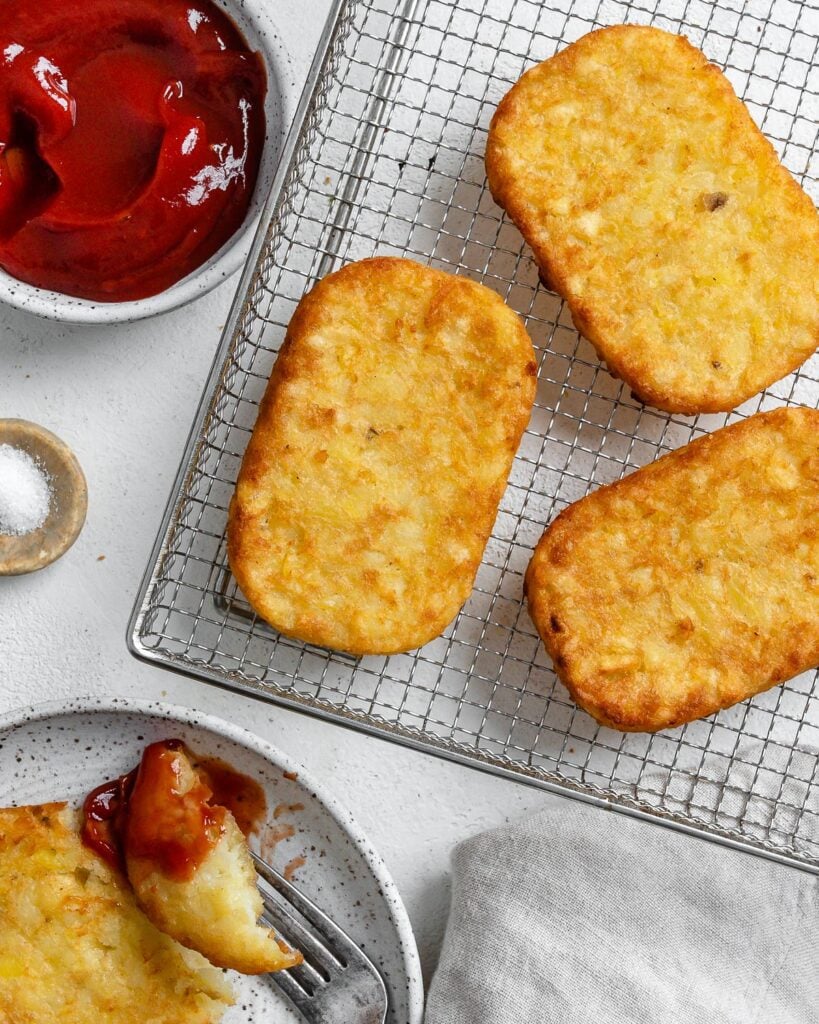 four completed hash brown patties on a air fryer tray with ketchup and salt in the background