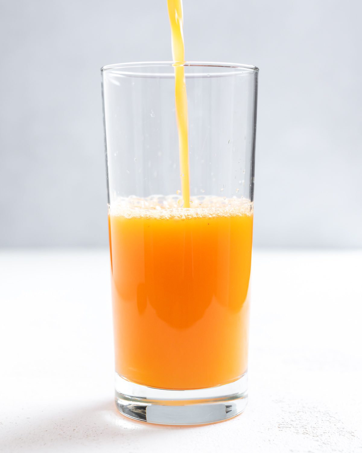Completed Apple OJ Carrot Juice being poured into a glass against a white surface and light background.