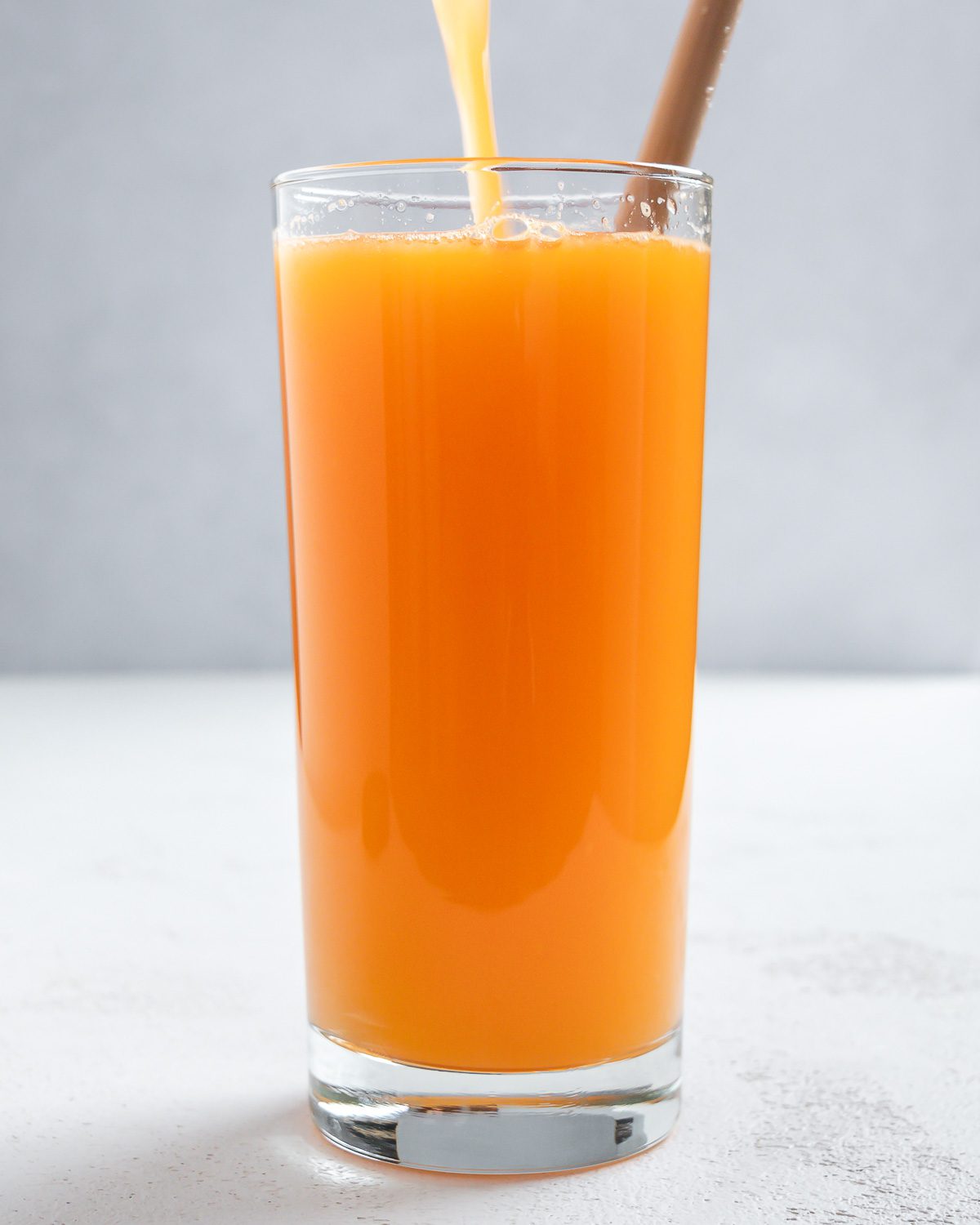 Completed Apple OJ Carrot Juice being poured into a glass against a white surface and light background.