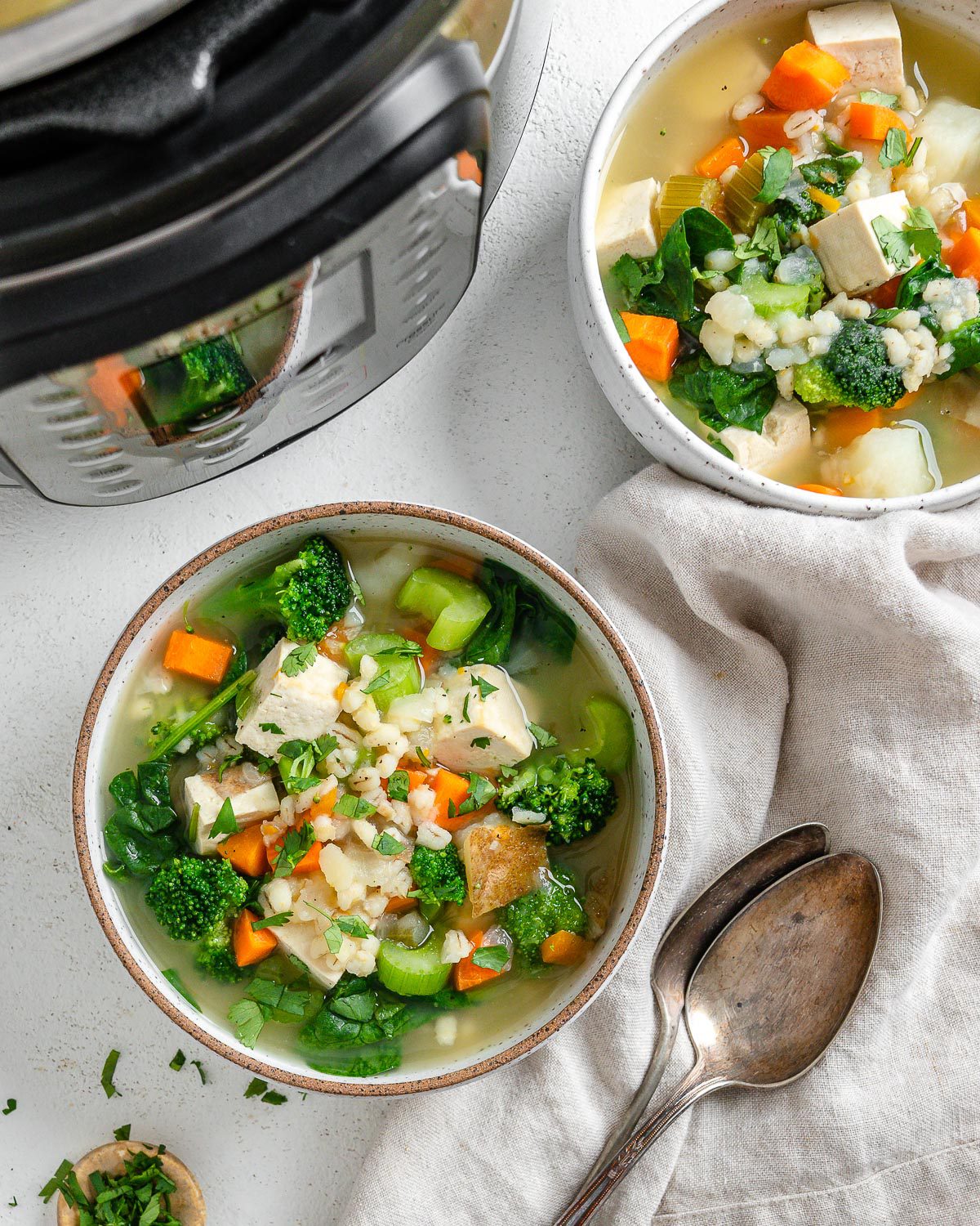 two bowls of completed Instant Pot Vegetable Barley Soup in a white bowl against a white background with an instant pot in the background