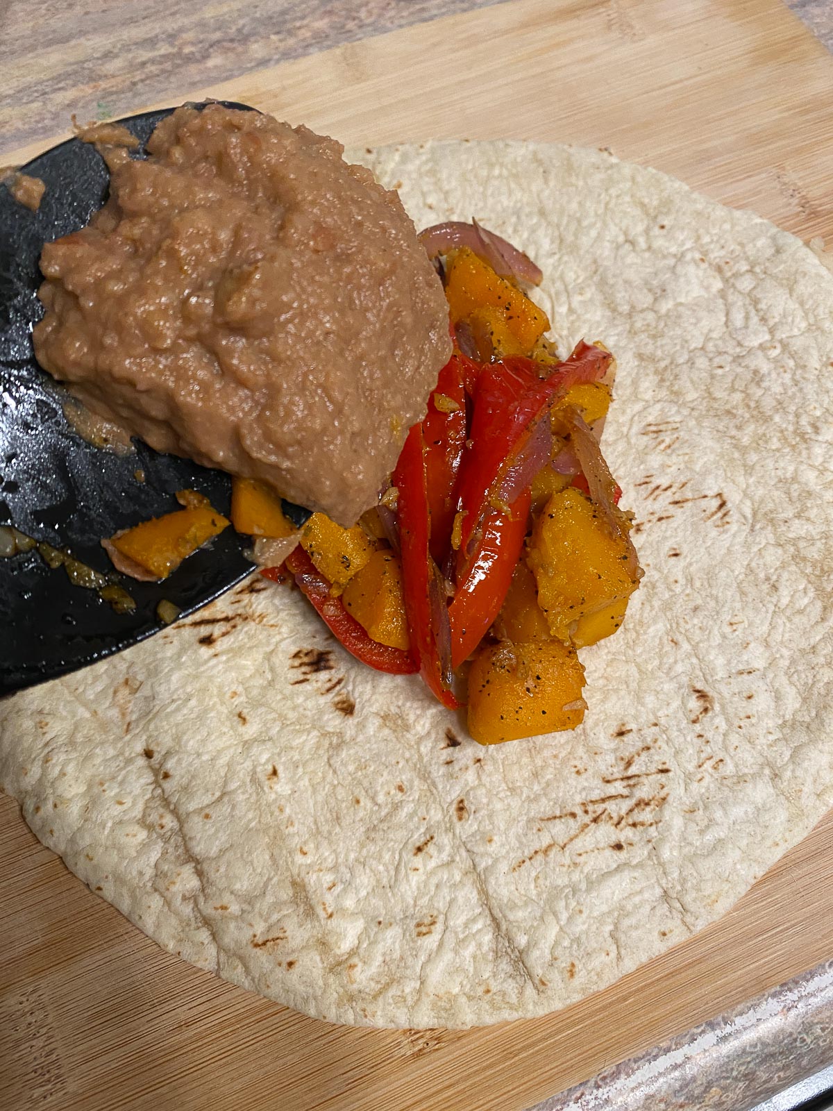 process of adding refried beans to butternut squash on a tortilla