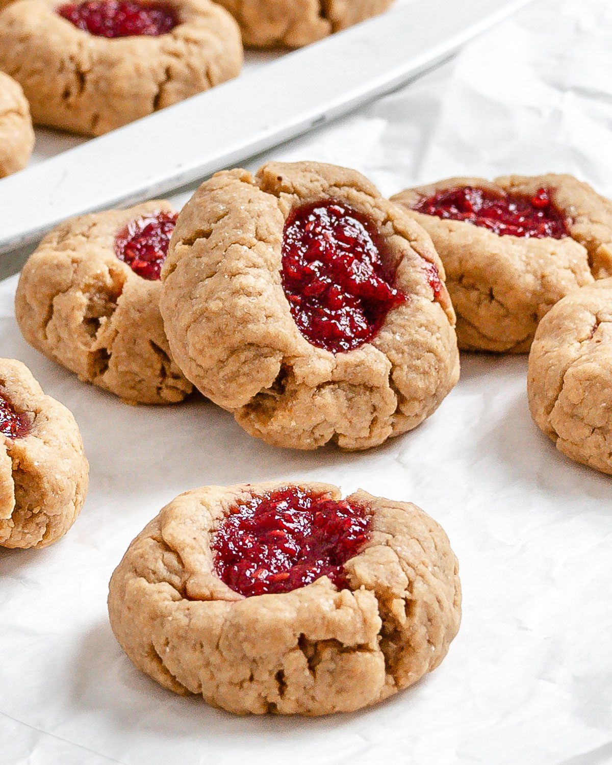 completed Vegan Peanut Butter and Jelly Thumbprint Cookies scattered on cookie sheet and white surface