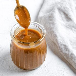 completed Simple Vegan Caramel in a jar with caramel drizzling down from a spoon against a white background