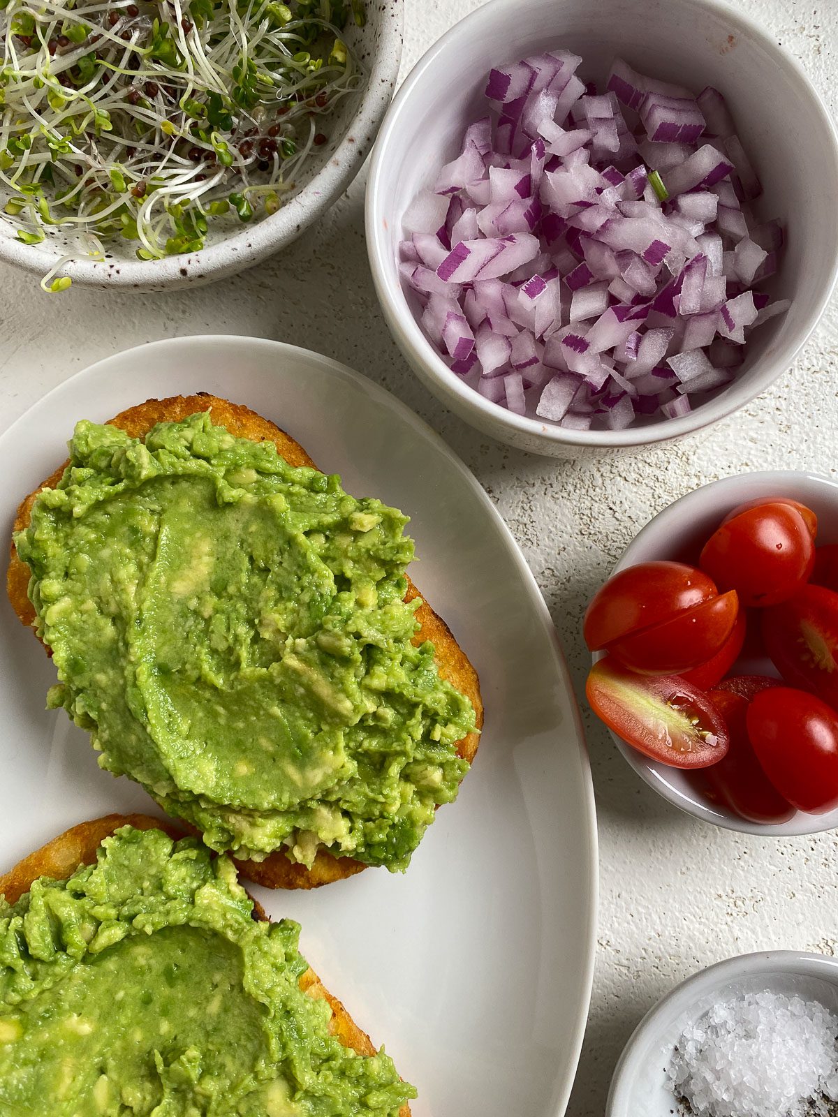 two slices of smashed avocado on hash browns alongside of toppings