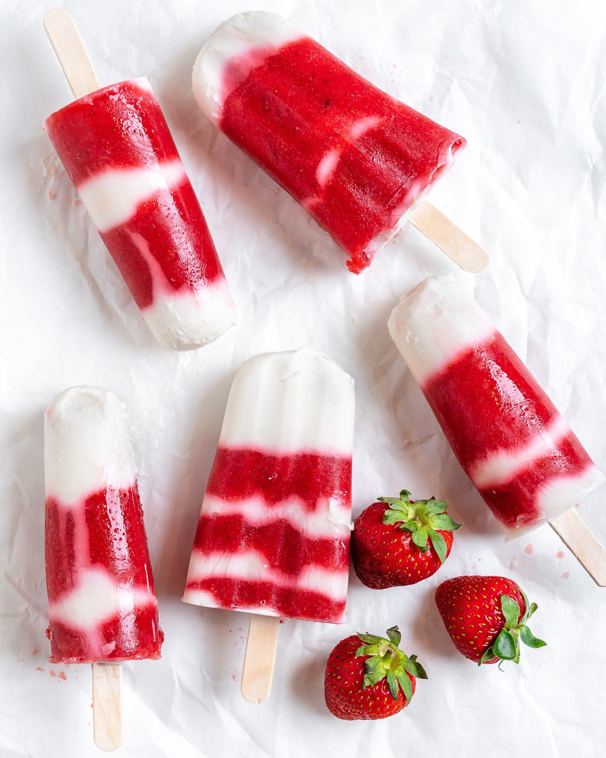 several completed Vegan Strawberry Popsicles on a white surface with strawberries alongside of them