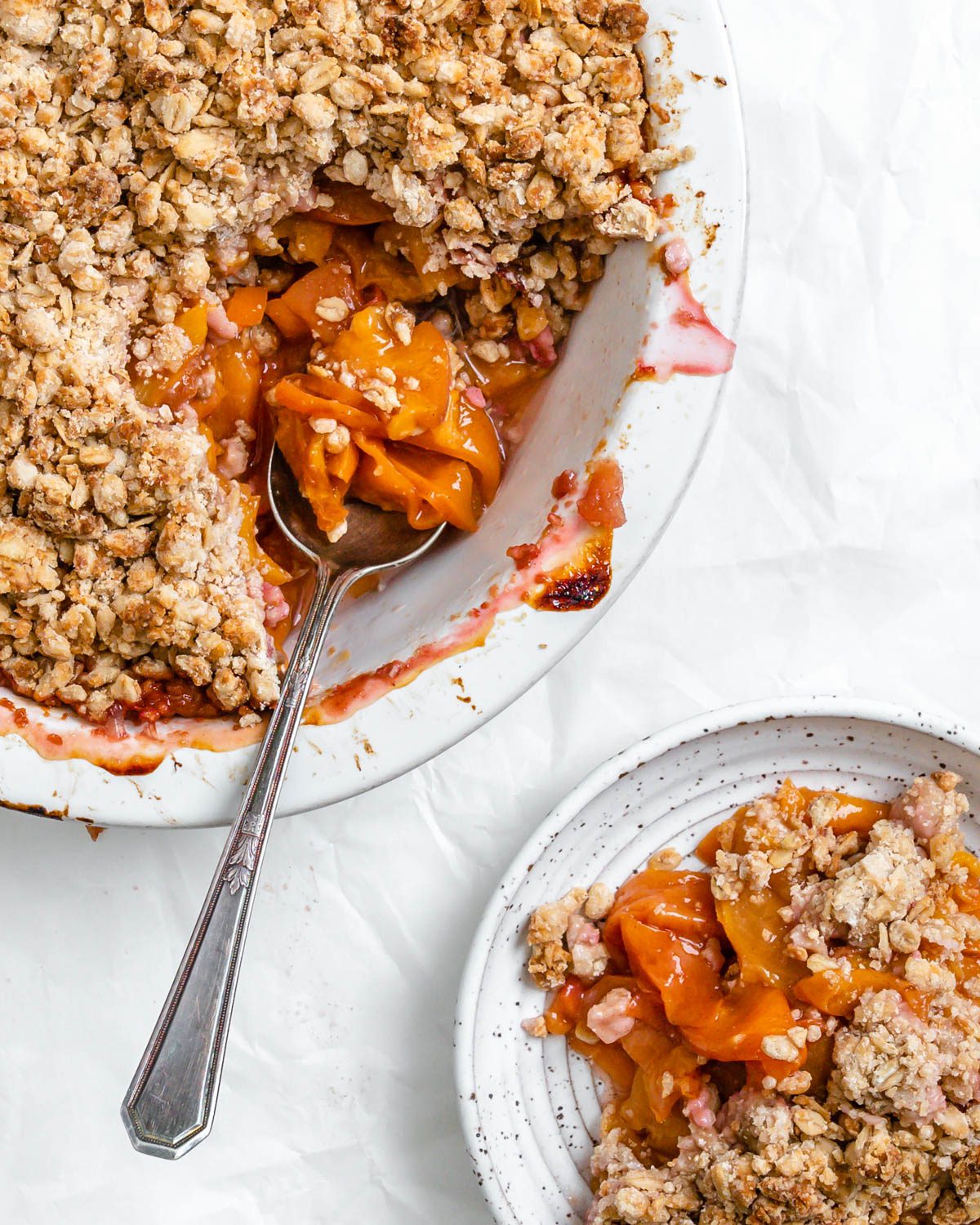 completed Vegan Peach Crisp in a white baking dish and with a piece plated on a plate against a white background