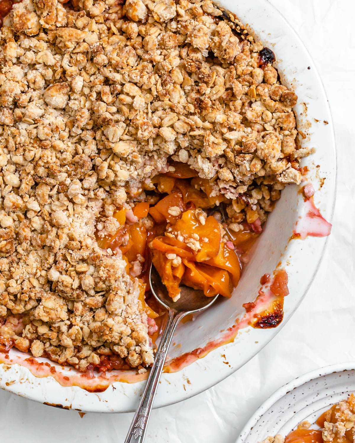completed Vegan Peach Crisp in a white baking dish and with a piece plated on a plate against a white background