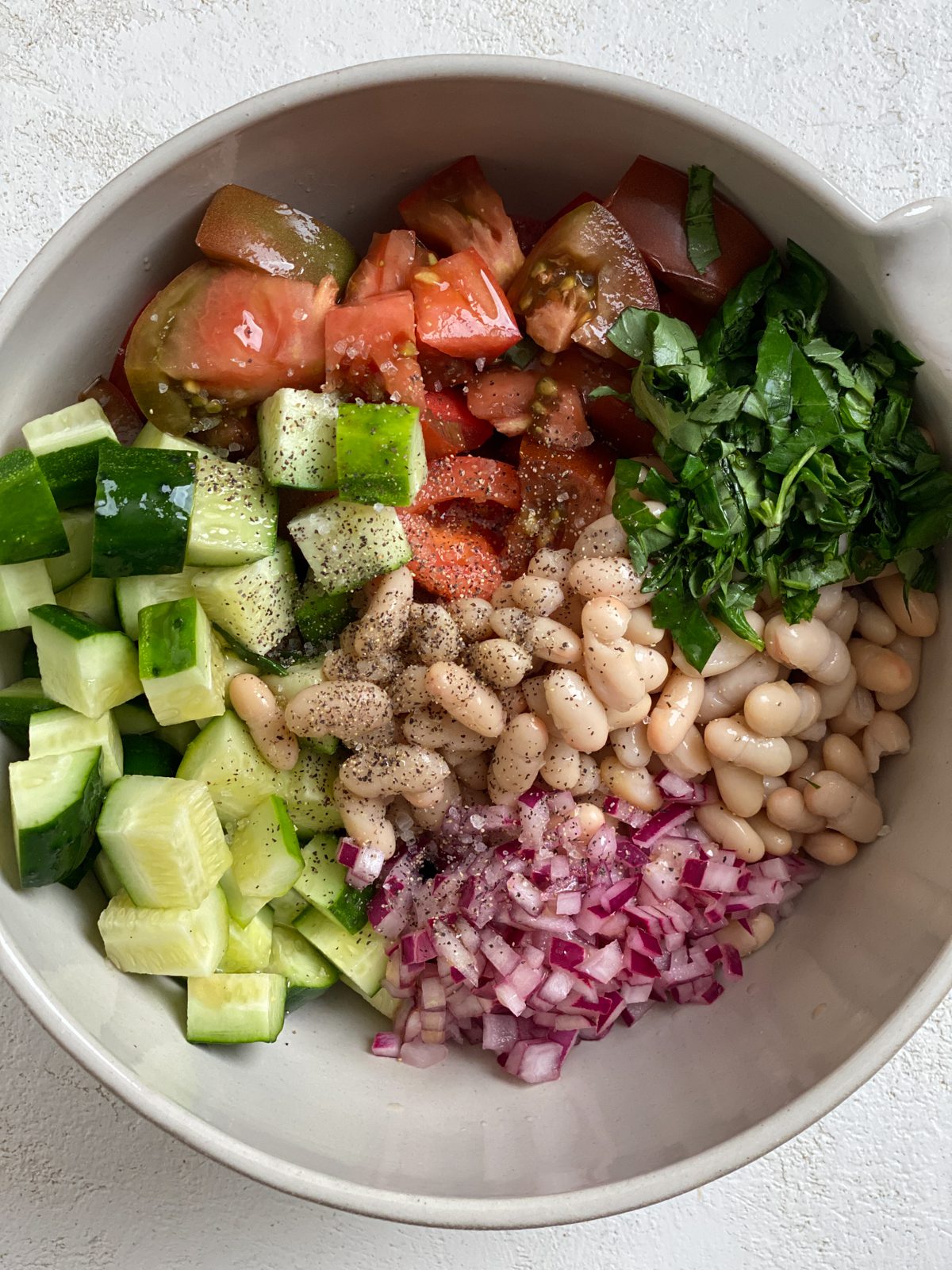 Process shot showing the addition of Easy Mediterranean White Bean Salad ingredients into a bowl.