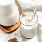 completed Super Creamy Vegan Eggnog in a glass alongside a jug of eggnog against a light surface