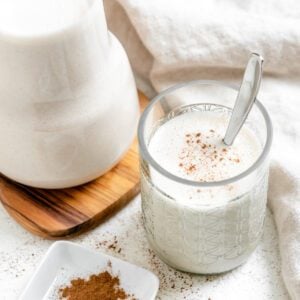 completed Super Creamy Vegan Eggnog in a glass alongside a jug of eggnog against a light surface
