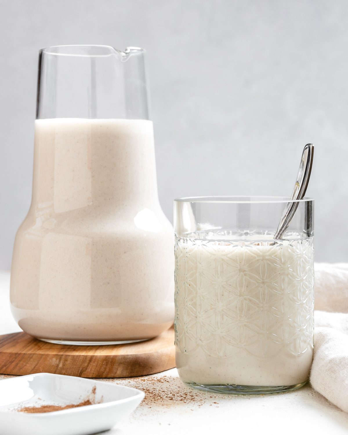 completed Super Creamy Vegan Eggnog in a glass alongside a jug of eggnog against a light surface