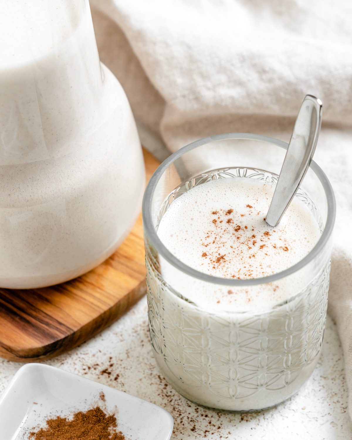 completed Super Creamy Vegan Eggnog in a glass alongside a jug of eggnog against a light surface