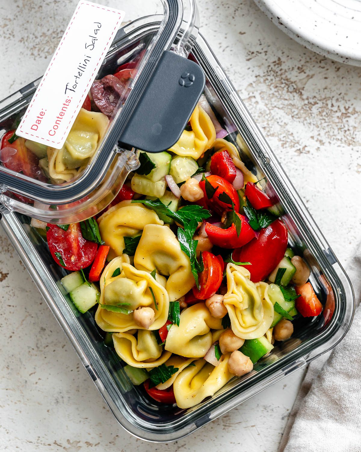 Tortellini pasta salad stored in a glass container. The lid of the container is off and to the side with a label on it.
